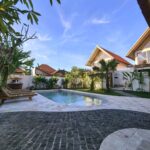 Daytime view of the outdoor swimming pool surrounded by wooden sunbeds, lush tropical gardens, and unique A-frame cabins at Kuda Putih Pandawa Guesthouse Bali.