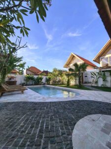 Daytime view of the outdoor swimming pool surrounded by wooden sunbeds, lush tropical gardens, and unique A-frame cabins at Kuda Putih Pandawa Guesthouse Bali.