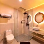 Modern ensuite bathroom featuring a glass walk-in shower with black rain shower fixtures and a backlit wooden mirror in the Deluxe Double room at Kuda Putih Pandawa.