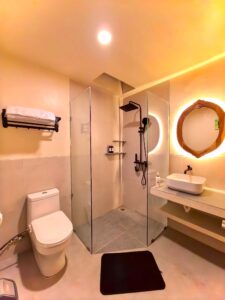 Modern ensuite bathroom featuring a glass walk-in shower with black rain shower fixtures and a backlit wooden mirror in the Deluxe Double room at Kuda Putih Pandawa.