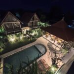 Elevated night view of the illuminated swimming pool, traditional dining pavilion, and tropical A-frame cabins at Kuda Putih Pandawa accommodation in Bali.
