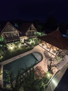 Elevated night view of the illuminated swimming pool, traditional dining pavilion, and tropical A-frame cabins at Kuda Putih Pandawa accommodation in Bali.