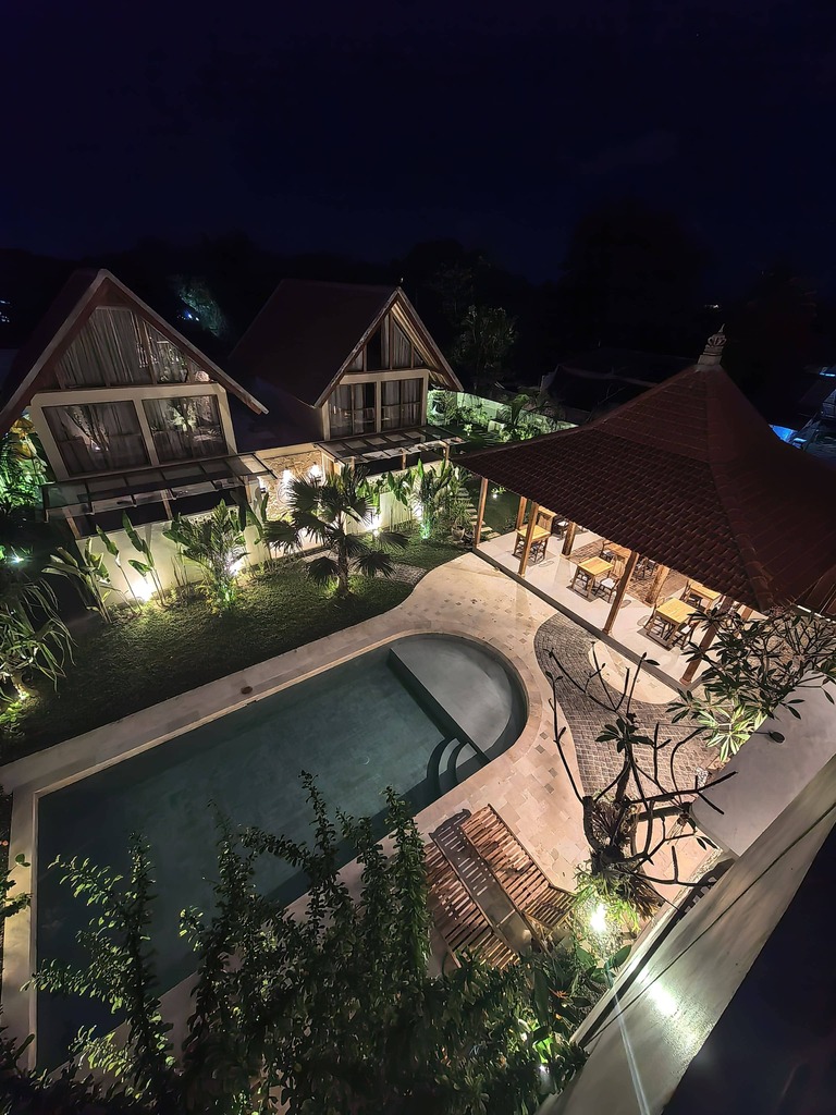 Elevated night view of the illuminated swimming pool, traditional dining pavilion, and tropical A-frame cabins at Kuda Putih Pandawa accommodation in Bali.
