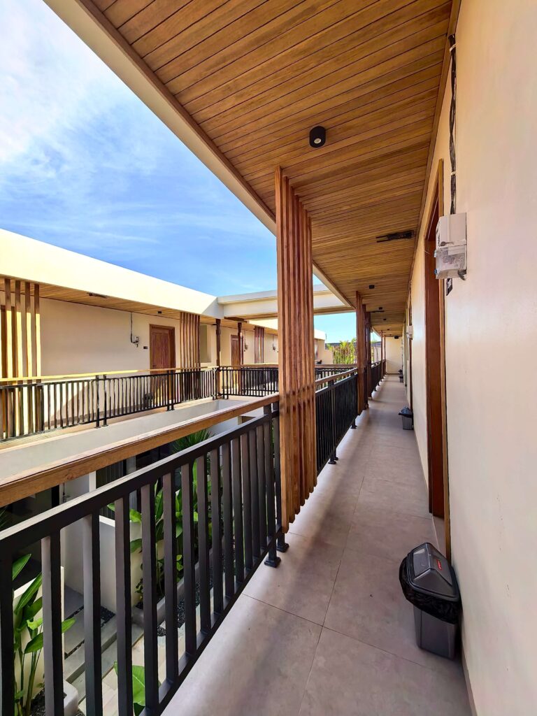Bright daytime atmosphere along the second-floor balcony walkway at Kuda Putih Pandawa Guesthouse Bali, showcasing wooden ceilings and guest rooms.