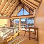 Sunlit wooden A-frame bedroom featuring a comfortable king bed, a wooden workspace, and large glass windows at Kuda Putih Pandawa premium guesthouse in Bali.