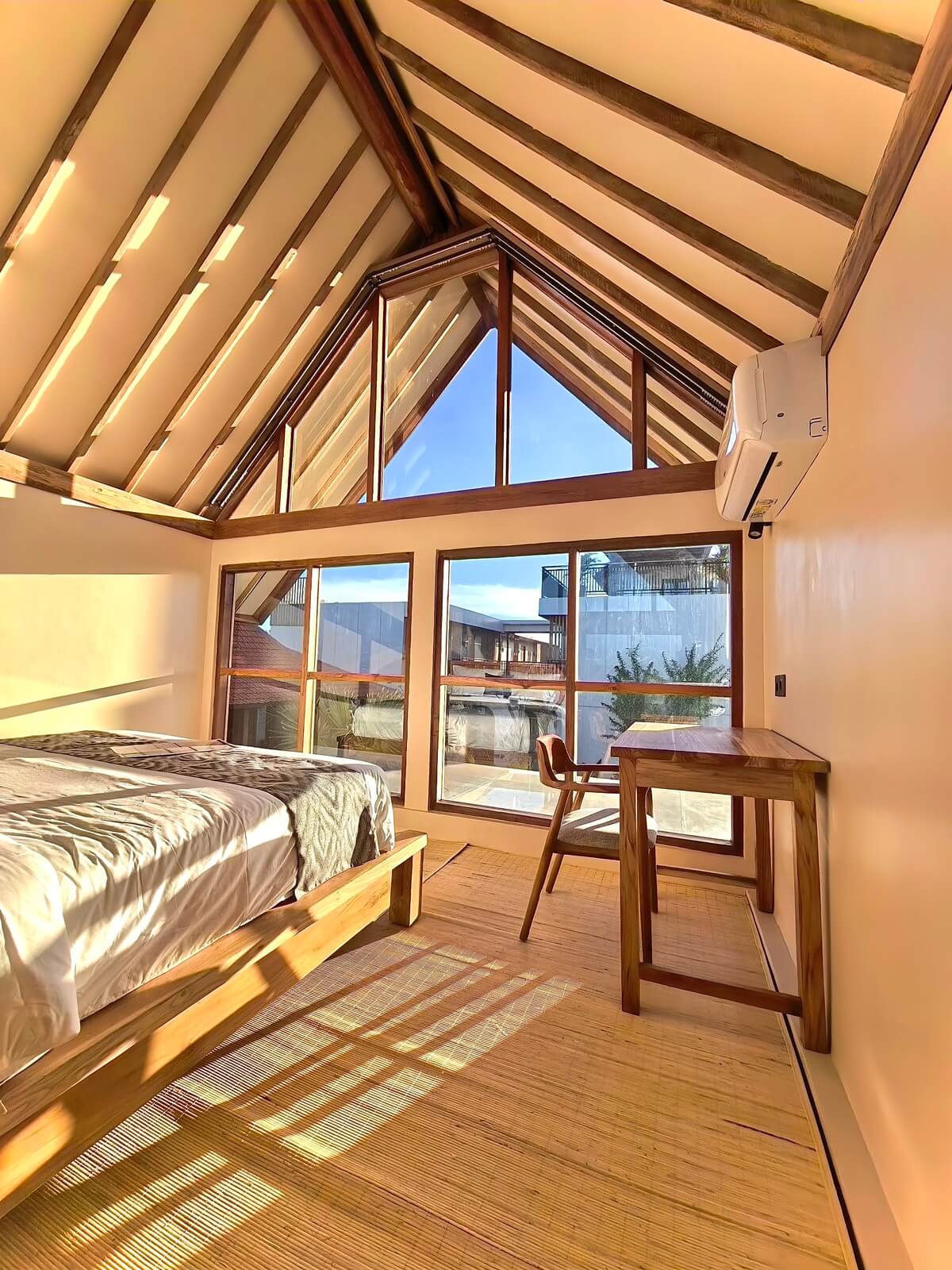 Sunlit wooden A-frame bedroom featuring a comfortable king bed, a wooden workspace, and large glass windows at Kuda Putih Pandawa premium guesthouse in Bali.