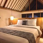 A warm and inviting bedroom at night under a wooden A-frame roof, featuring a plush bed with a branded Kuda Putih pillow and rattan lighting in Bali.