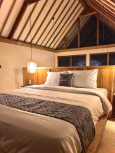 A warm and inviting bedroom at night under a wooden A-frame roof, featuring a plush bed with a branded Kuda Putih pillow and rattan lighting in Bali.