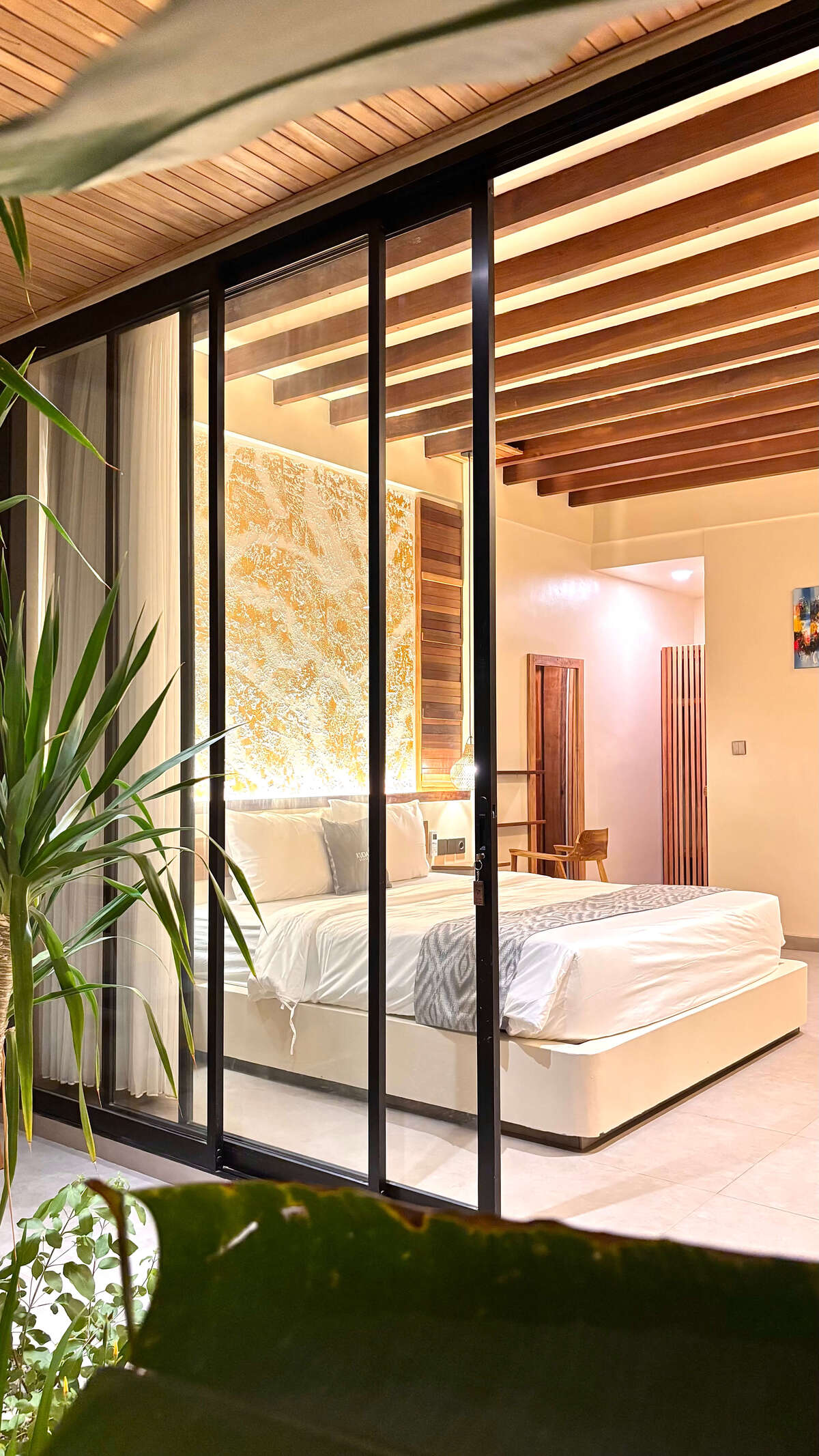 A view through glass sliding doors into the ground-floor Deluxe Double room at Kuda Putih Pandawa guesthouse in Bali, featuring a cozy bed and warm aesthetic lighting.