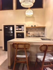 A fully equipped modern kitchenette with a dining island, rattan bar stools, and warm pendant lighting inside a premium suite at Kuda Putih Pandawa South Kuta.