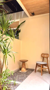 A tranquil private garden patio featuring a wooden chair, side table, and tropical plants outside the Deluxe Double room at Kuda Putih Pandawa South Kuta.