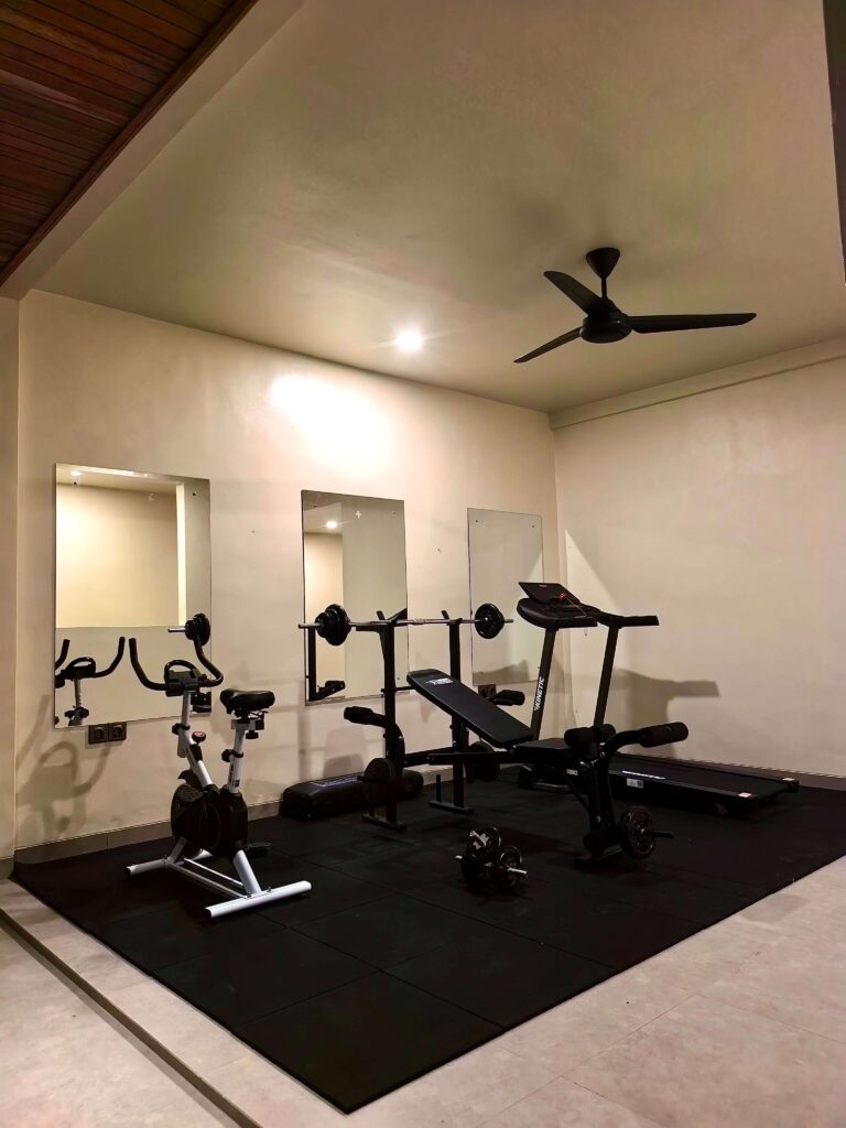 A well-equipped indoor private gym at Kuda Putih Pandawa guesthouse in Bali, featuring a stationary bike, weight bench, treadmill, and wall mirrors on a rubber mat flooring.