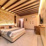 A wide interior view of the spacious Deluxe Double room at Kuda Putih Pandawa Bali, showcasing an illuminated textured headboard, wooden ceiling beams, and a king-size bed.