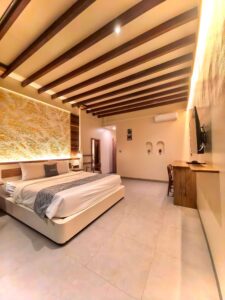 A wide interior view of the spacious Deluxe Double room at Kuda Putih Pandawa Bali, showcasing an illuminated textured headboard, wooden ceiling beams, and a king-size bed.